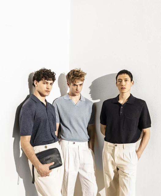 Three adult men stand in a photo studio wearing polo shirts and light-colored pants. One man holds a black clutch. Studio lighting.