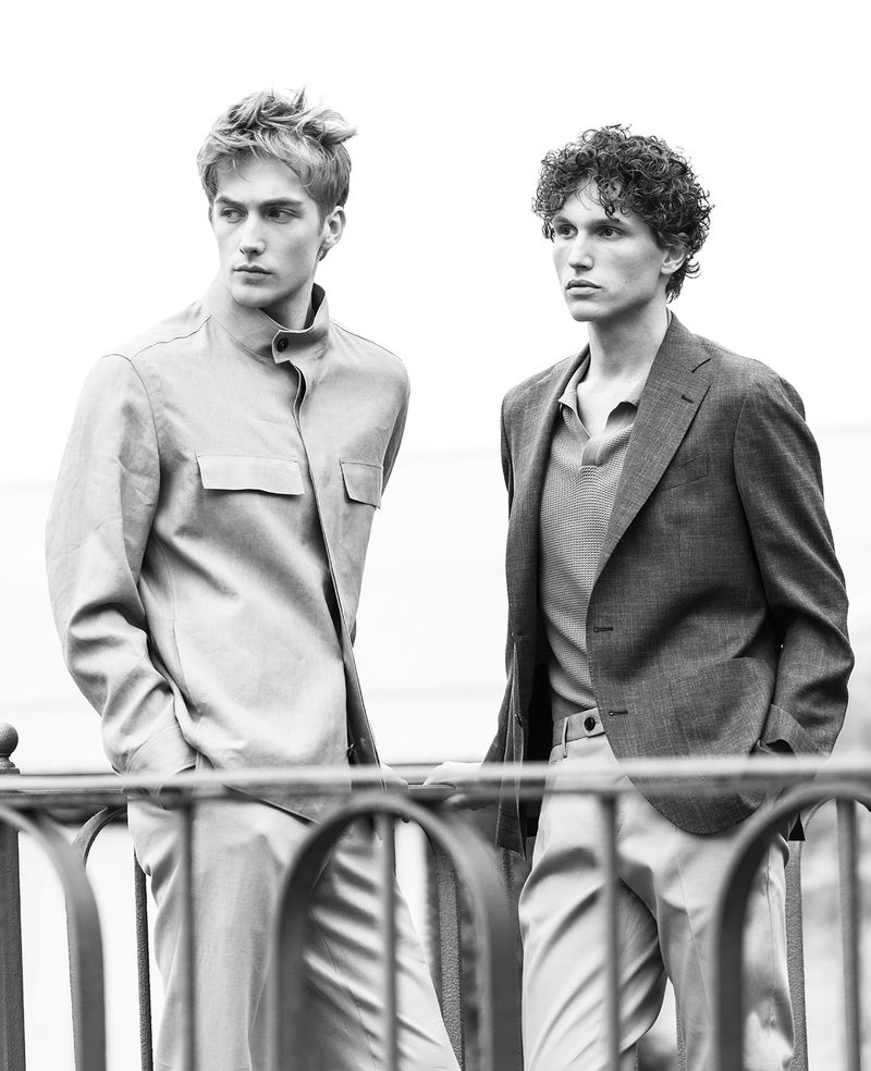 Black and white portrait of two young adult males wearing jackets and dress pants, looking off into the distance on a cloudy day.