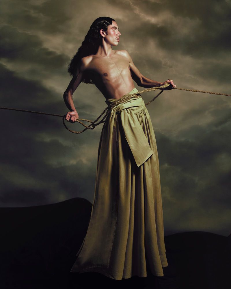 Topless male model in long skirt, holding rope, posing against a backdrop of dark storm clouds and ground.