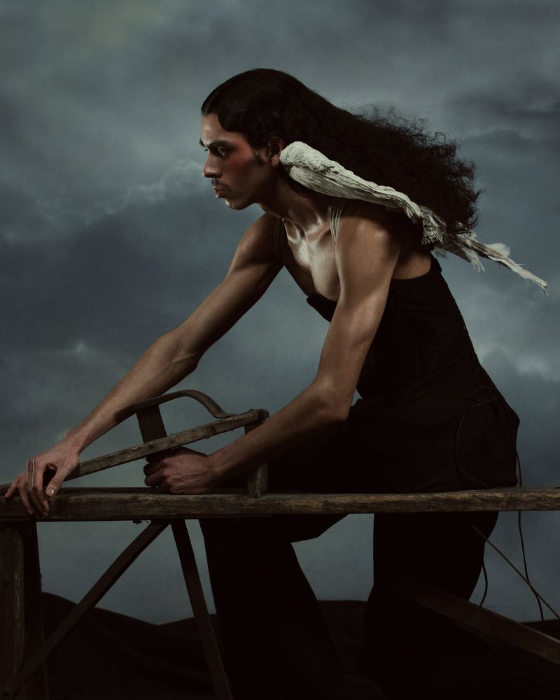 Portrait of a man with long dark hair wearing a black tank top and trousers sitting on a wooden sawhorse with a cloudy background.