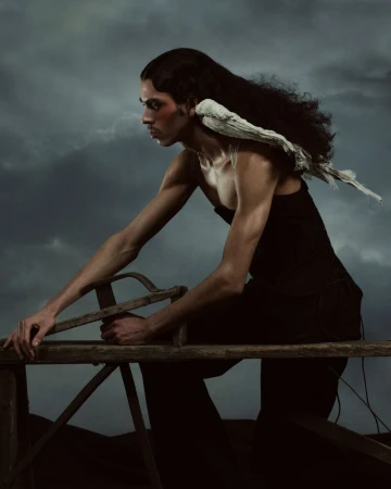 Portrait of a man with long dark hair wearing a black tank top and trousers sitting on a wooden sawhorse with a cloudy background.