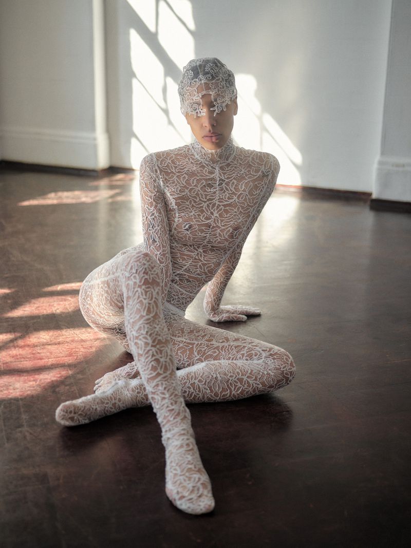 Fashionable woman poses in a white lace bodysuit indoors, with sunlight streaming in from windows on the white wall, accentuating the floor.