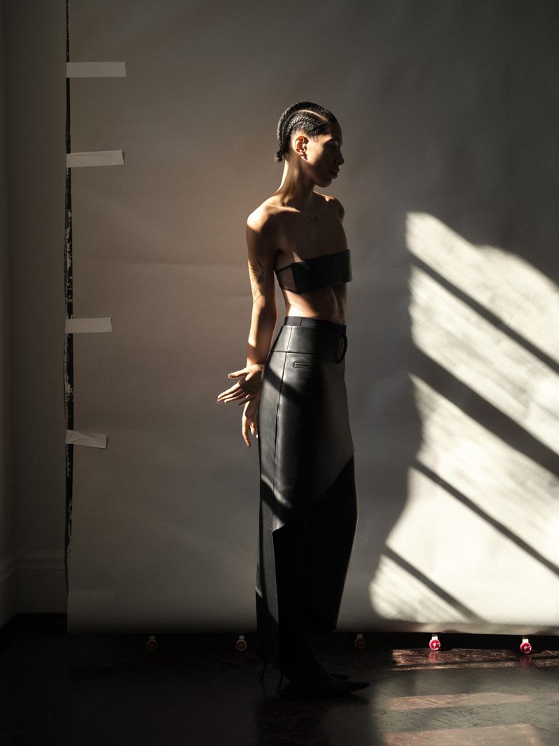 Fashionable non-binary person poses in a modern photo studio wearing a black leather skirt and bra top, bathed in bright directional light.