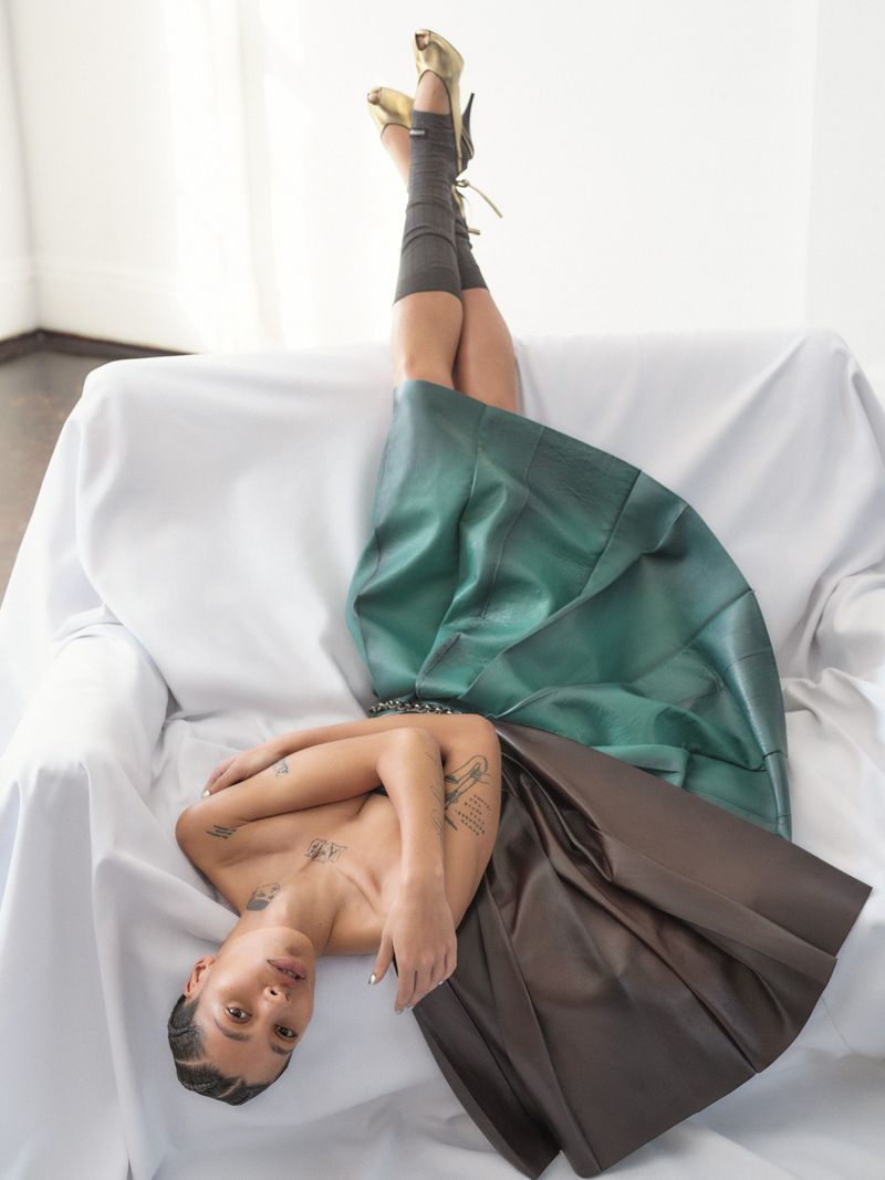 Woman lying on a white couch wearing a brown and green skirt and gold high heels in a light indoor setting.