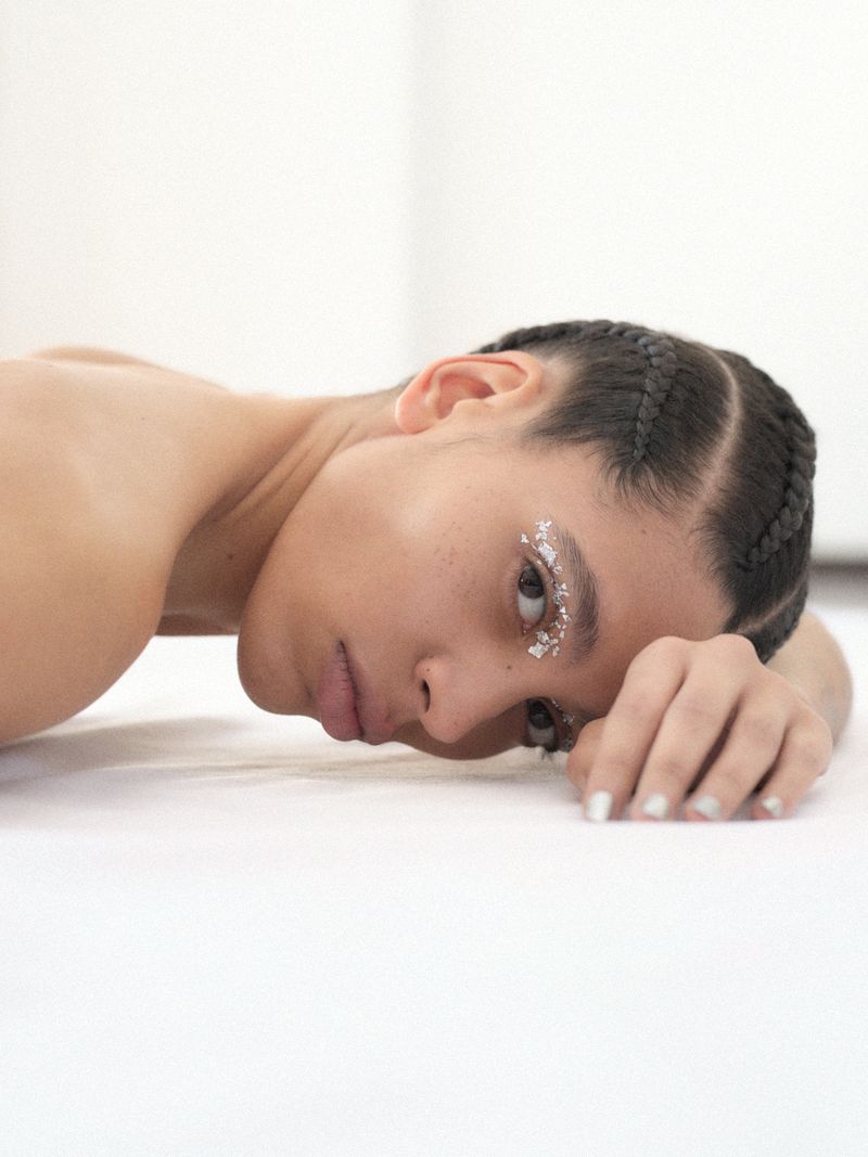 Adult female with braided hair and silver eye makeup lying down on white fabric in a light-filled indoor setting. Close up, portrait orientation.