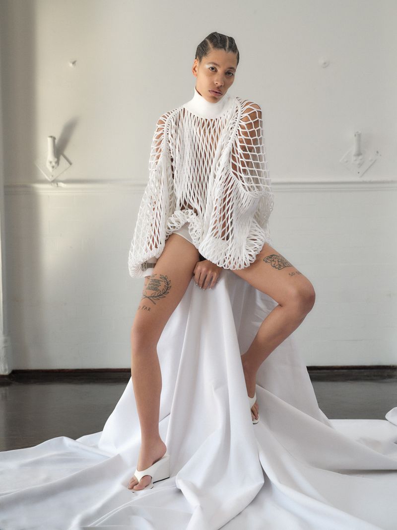 A young adult female in a white crocheted sweater sits on a draped white fabric, wearing white sandals, in a minimal, light-filled indoor space.