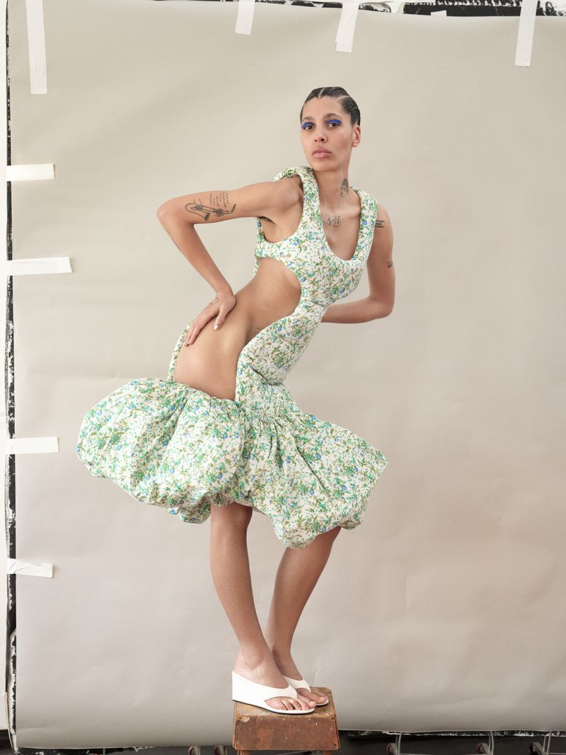 Woman poses in floral cut-out dress and white platform sandals on a wooden block against a neutral backdrop with white tape.
