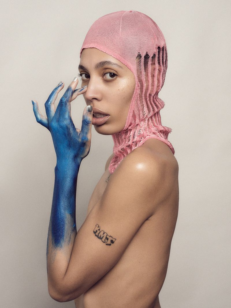 Person with blue painted arm and pink knitted headwear posing in a studio, portrait, fashion forward concept, beauty shot, neutral background.