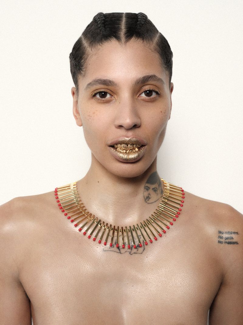 Striking portrait of a shirtless individual with gold grill teeth, braided hair, and a unique matchstick necklace against a white backdrop.