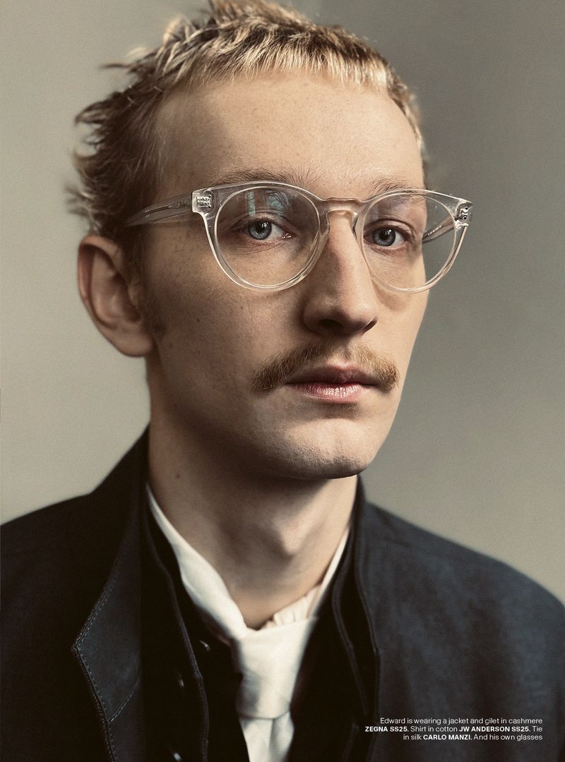 Close-up portrait of a blond adult male with a moustache wearing clear-framed glasses, a white shirt, and a dark blazer.