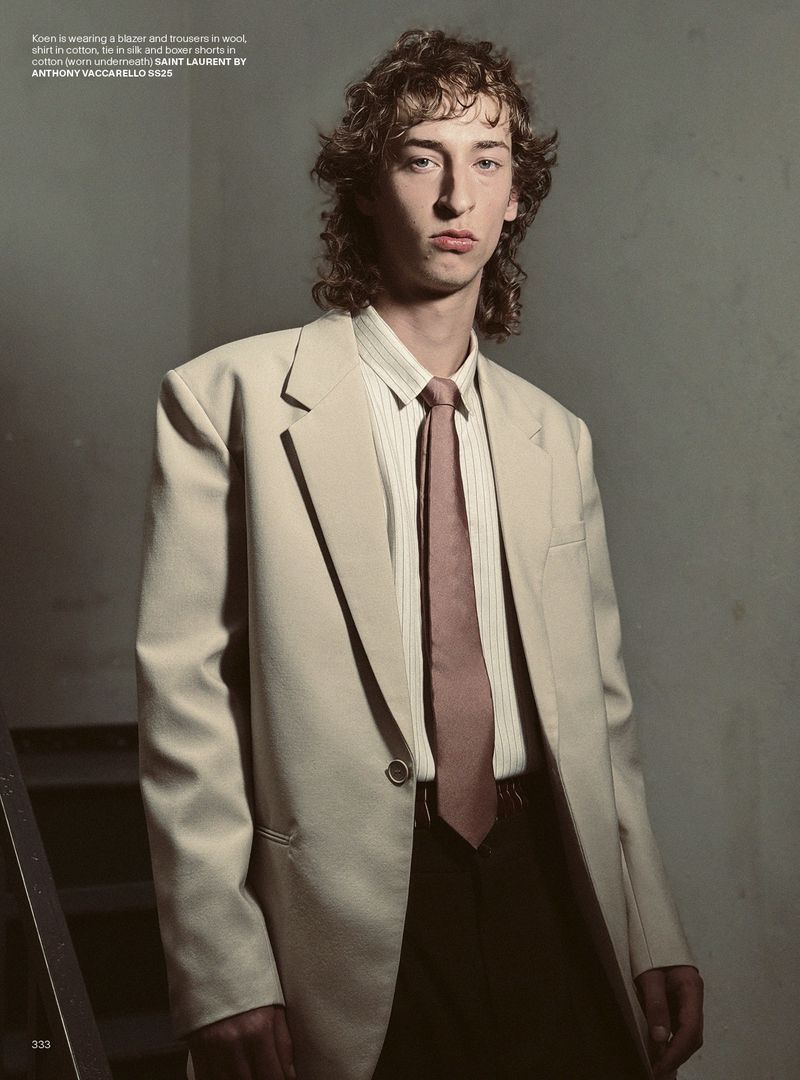 Portrait of a young male model in a beige wool blazer, striped shirt, and tie, posing indoors with dark background in a fashion editorial.