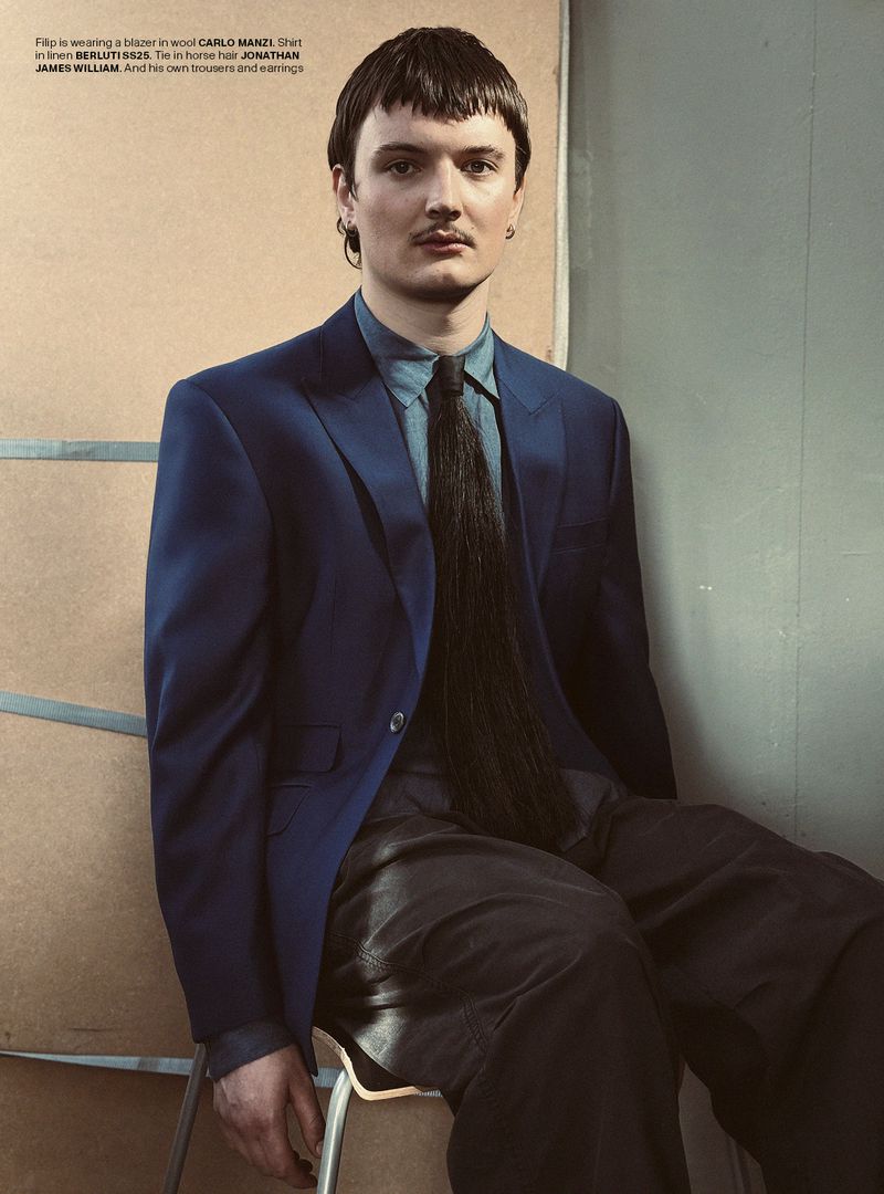 Male fashion model in blue suit, light blue shirt, and horsehair tie sits on a chair in a studio with natural light. Editorial fashion.