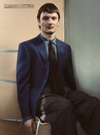 Male fashion model in blue suit, light blue shirt, and horsehair tie sits on a chair in a studio with natural light. Editorial fashion.
