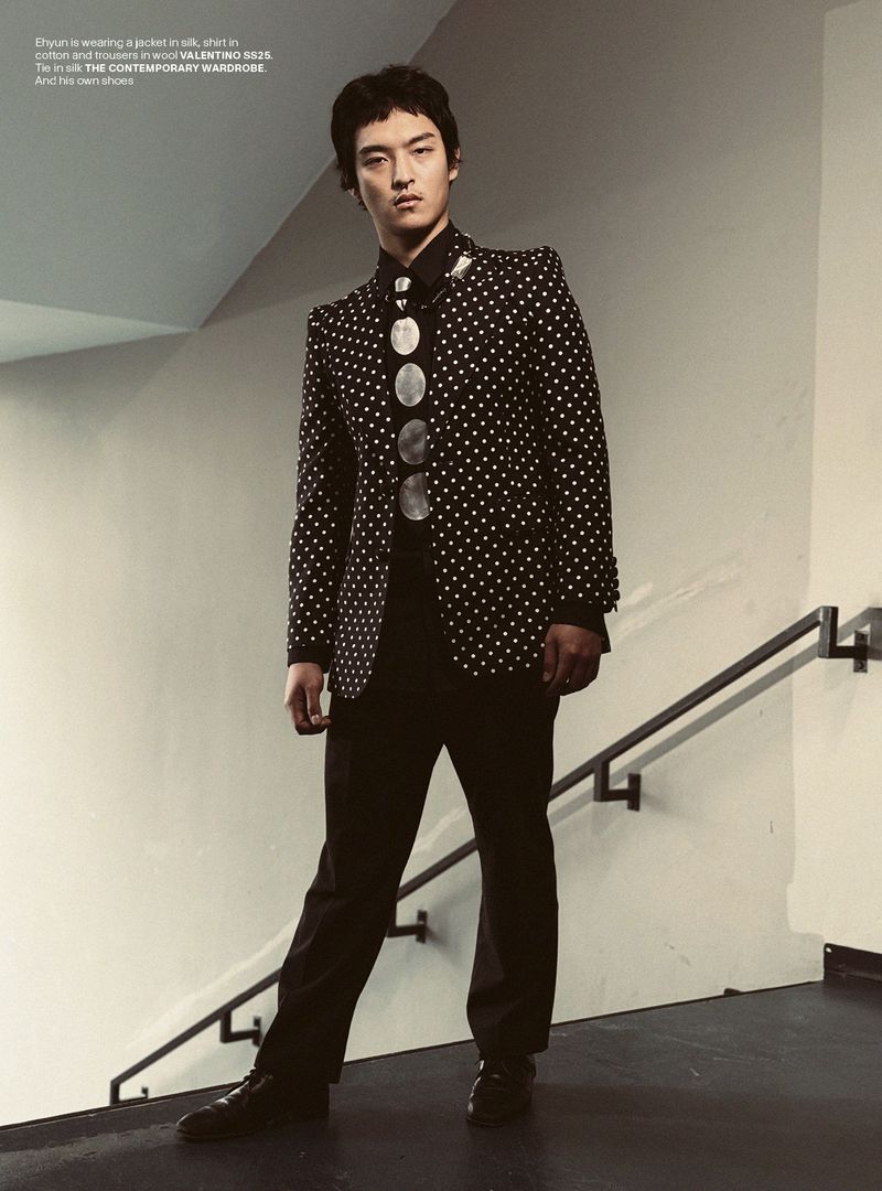 A stylish Asian man in a black and white jacket with black pants poses on a stairway with one hand in his pocket, looking forward.