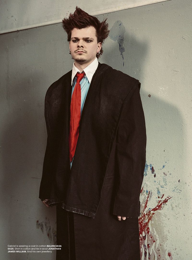 Stylish man in a dark jacket, light blue shirt, and red tie poses in a gritty indoor setting with paint splatters on the wall.
