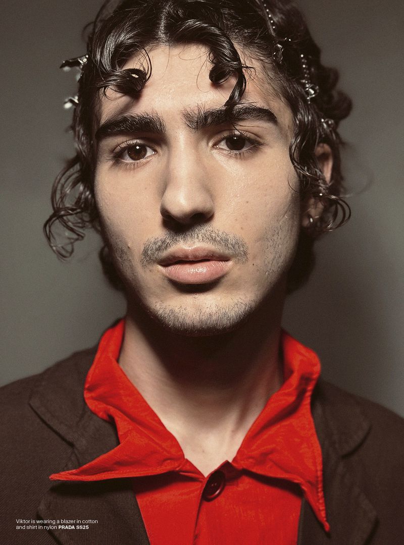 Close-up portrait of a young adult male with curly hair and a red shirt with black blazer, posing in front of a grey studio backdrop.