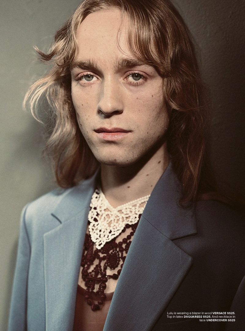 Portrait of a young adult with long blond hair, wearing a blue jacket and a white and black lace collar, posing in a studio setting.