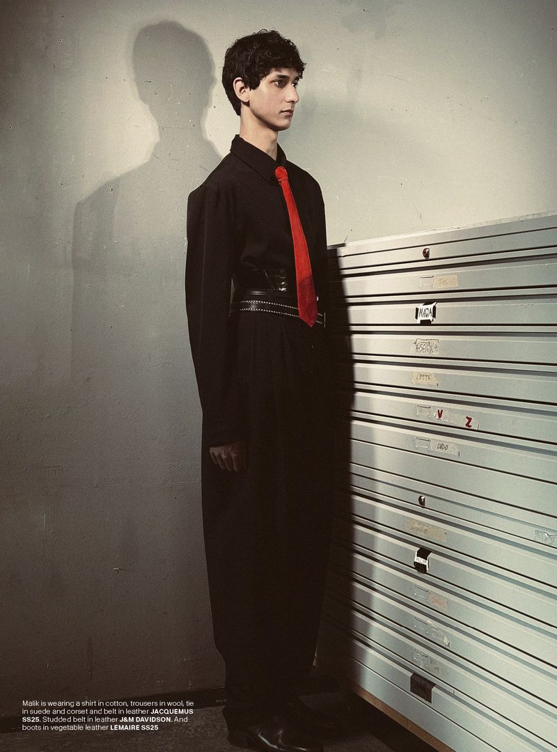 Fashion editorial shot of a young adult male in a black outfit and bright red tie, posing next to metal filing drawers in studio.
