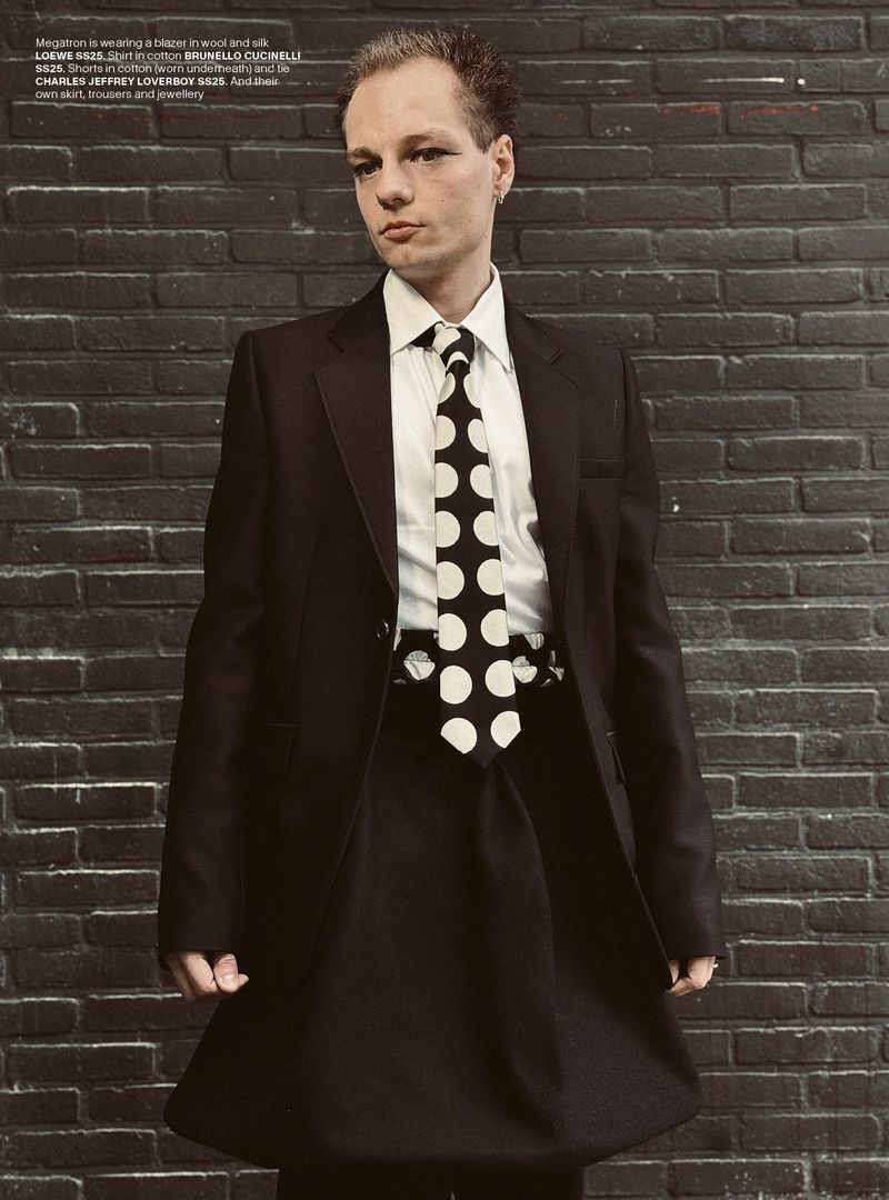Person in dark suit and polka dot tie standing against a dark brick wall, fashionably posing in formal attire. Style and confidence.
