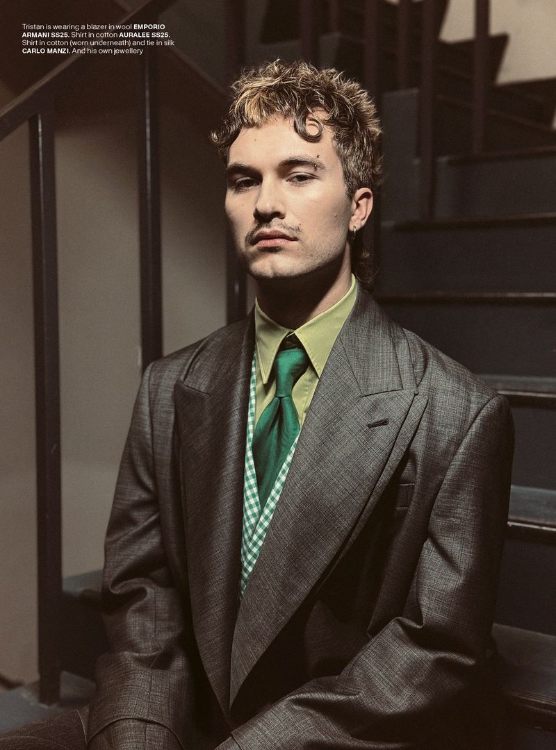 Portrait of a handsome man in a grey suit, green shirt, and matching tie, sitting on dark stairs in an indoor setting. Fashion editorial.