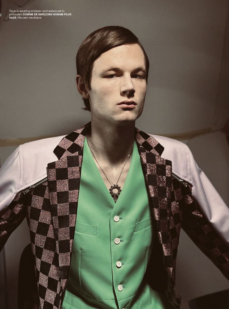 Man in green vest and patterned blazer poses indoors. He wears a pendant necklace and has dark hair styled with a side part.