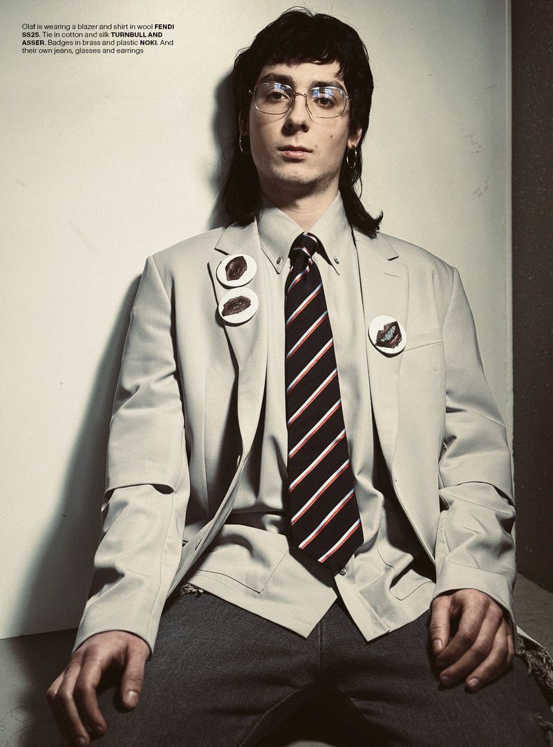 Portrait of a young adult male with glasses wearing a blazer, striped tie, and grey pants, in a photo studio setting with neutral colors.