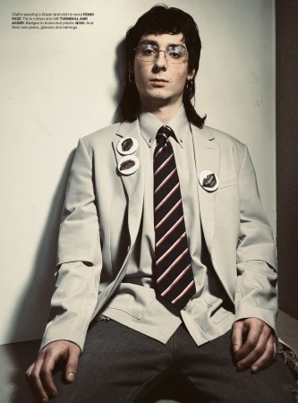 Portrait of a young adult male with glasses wearing a blazer, striped tie, and grey pants, in a photo studio setting with neutral colors.