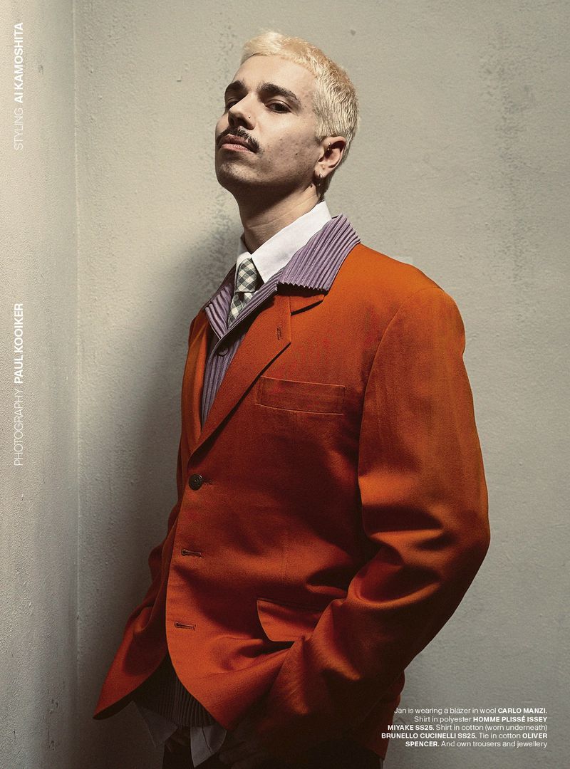 Stylish portrait of a blond male in a red blazer, collared shirt, and tie, posed in a corner with neutral background, fashion editorial.