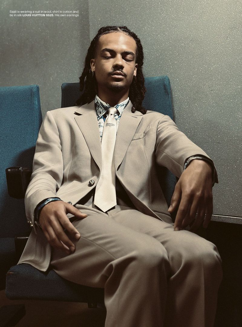 Portrait of a man wearing a suit and tie, seated in a chair with his eyes closed, illuminated by soft, ambient light in an indoor setting.
