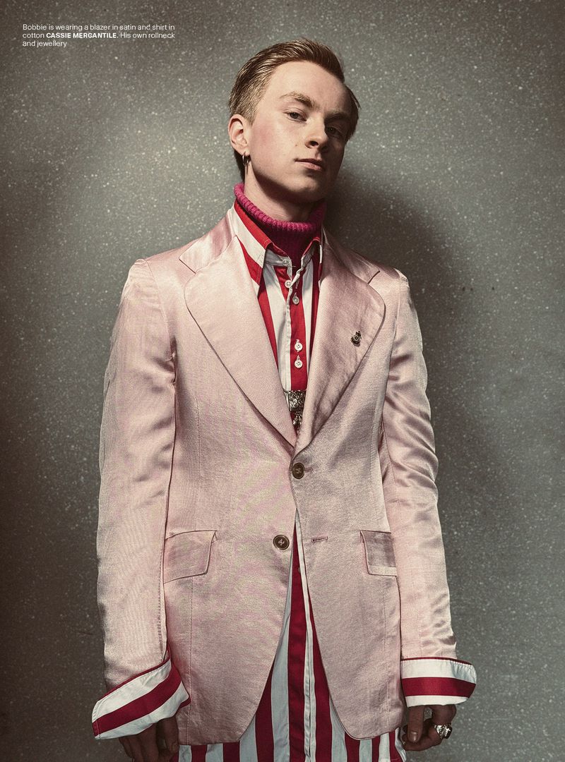 Portrait of a fair-skinned male model in a light pink suit with a red turtleneck and striped shirt against a grey textured backdrop.