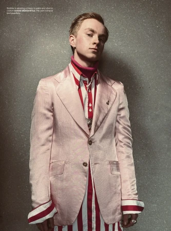Portrait of a fair-skinned male model in a light pink suit with a red turtleneck and striped shirt against a grey textured backdrop.