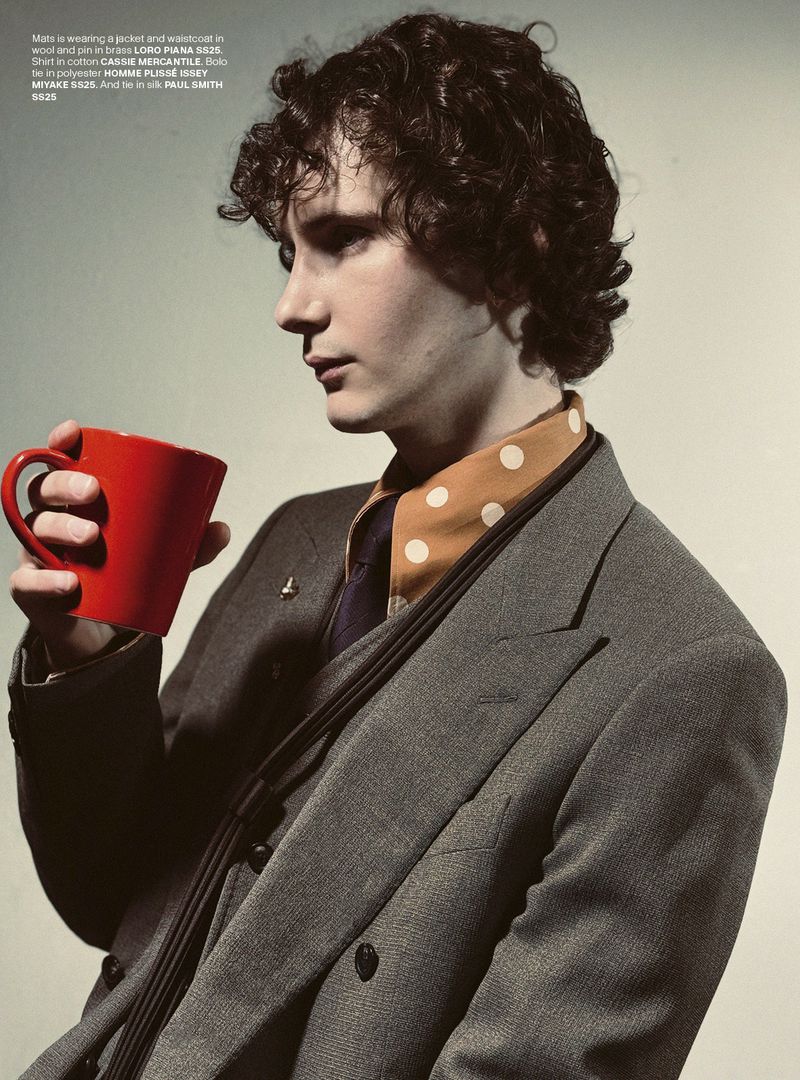 Young man in a suit holds a red mug, with a patterned scarf showing at the neck. Indoor portrait with a neutral backdrop.