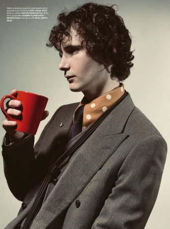 Young man in a suit holds a red mug, with a patterned scarf showing at the neck. Indoor portrait with a neutral backdrop.