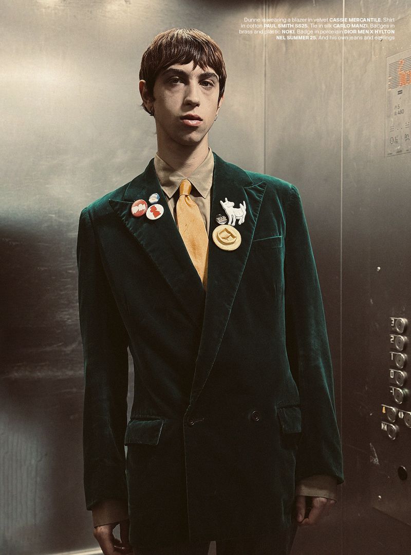 A young adult stands in an elevator wearing a dark green velvet suit, yellow tie, and several pins on the lapel, looking at camera.