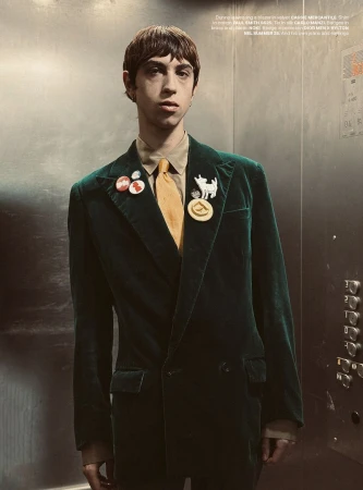 A young adult stands in an elevator wearing a dark green velvet suit, yellow tie, and several pins on the lapel, looking at camera.