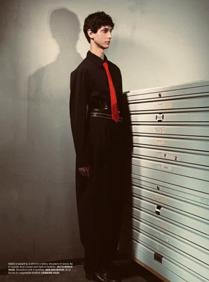 Young man in black shirt and trousers with a red tie standing beside a large metallic storage unit in an editorial portrait.