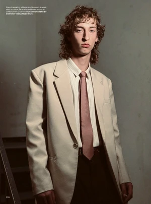 Young man with curly brown hair wearing a beige blazer, striped shirt, and tie against a grey wall