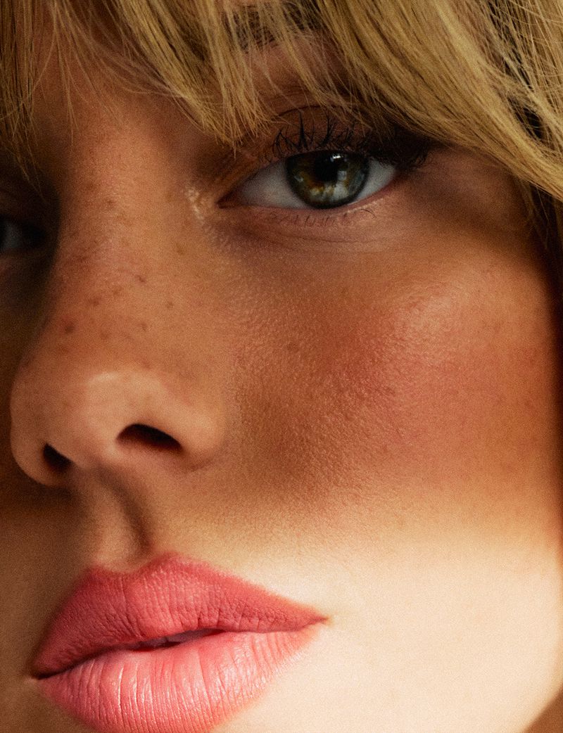 Close-up portrait of a blonde adult woman with fair skin, freckles, and green eyes. She has pink lips and wears subtle makeup.