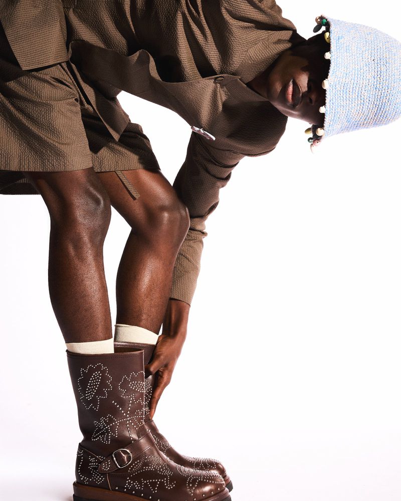 Dark-skinned model wearing brown suit, light blue hat, and embroidered cowboy boots posing in a contemporary fashion editorial.