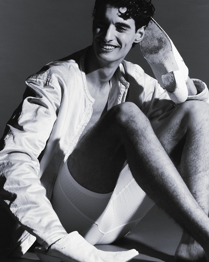 A man with curly dark hair sits shirtless, wearing white bottoms and a jacket, one foot in the air, smiling in a studio shot.