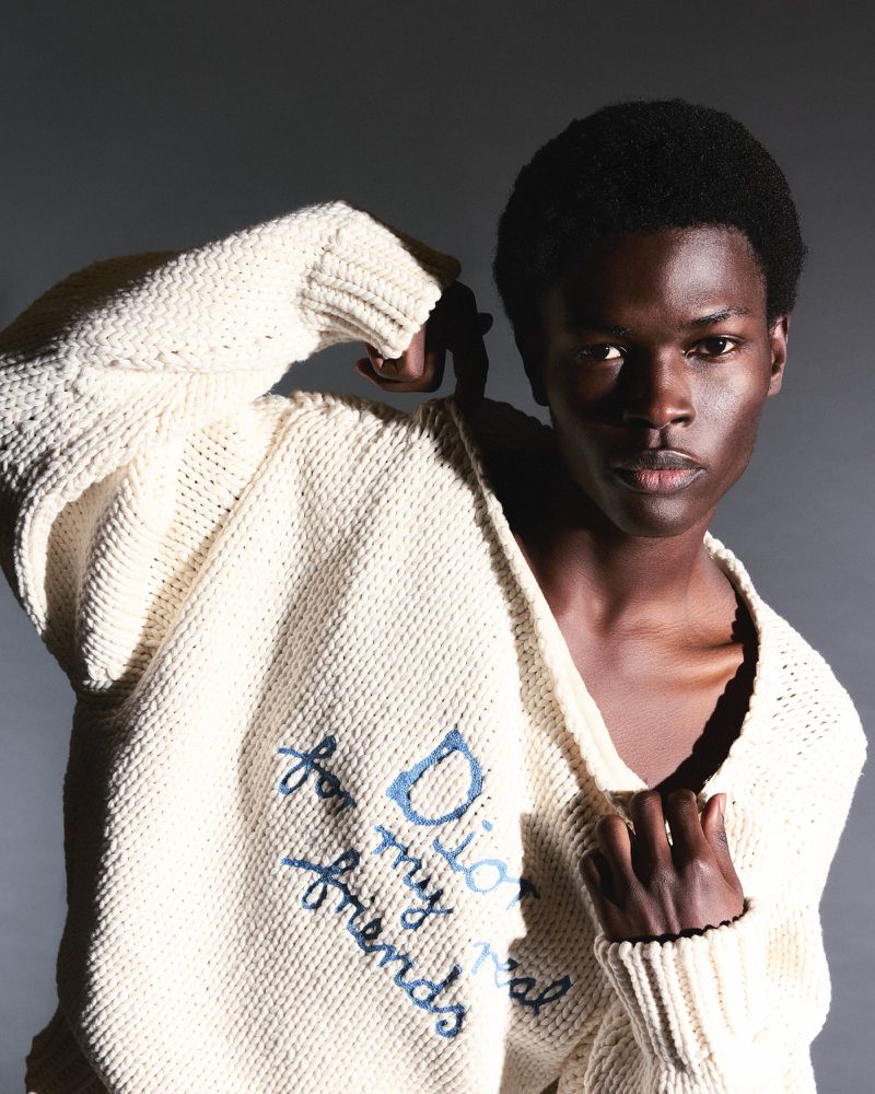 Close-up of Black male fashion model in off-white knit sweater with blue embroidery in studio against grey background. Fashion editorial.