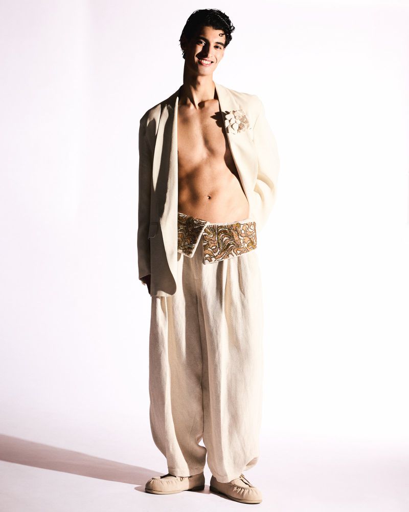 A young man strikes a pose in a relaxed suit, complemented by beige shoes, on a bright white backdrop. The outfit is fashion-forward.