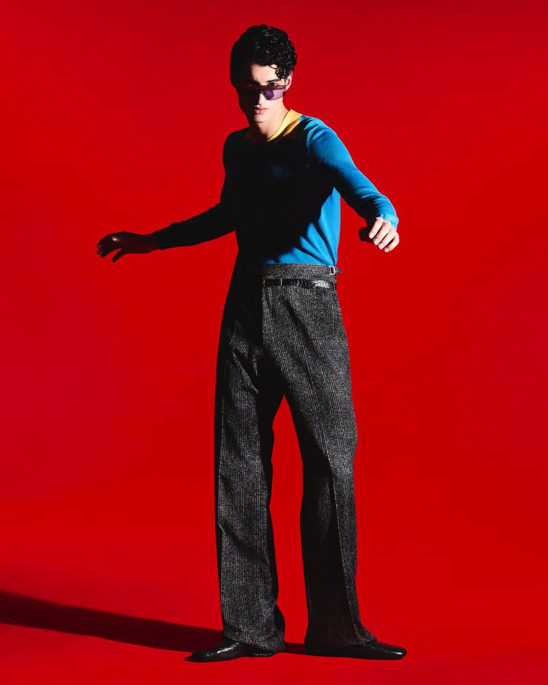 Adult male with dark curly hair in sunglasses and retro fashion poses in front of a seamless bright red background in a studio.