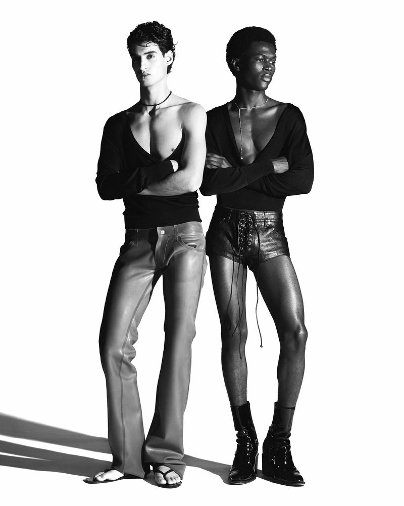 Black and white studio fashion shot of two young adult male models wearing low-cut tops, pants, shorts, and boots with arms crossed.