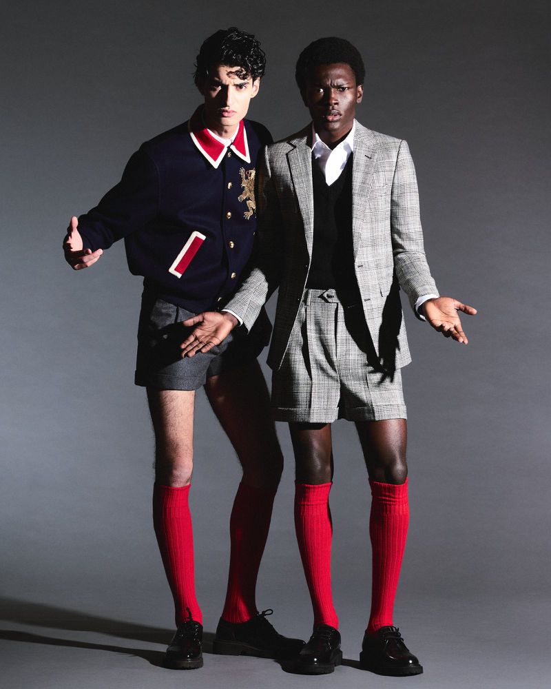 Two fashion models pose in matching grey shorts suits and red socks against a gray background, looking directly at the camera.