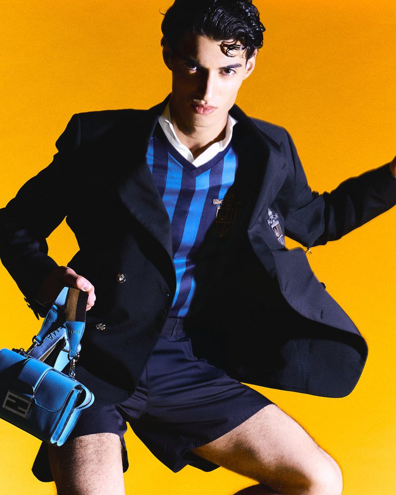 Male fashion model poses in blue blazer, shorts, and a striped sweater against a solid yellow backdrop in a studio setting.