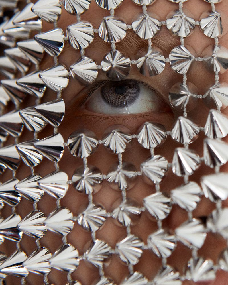Close-up of one eye of a person wearing a silver spiked decorative face covering, with a neutral expression and white background.