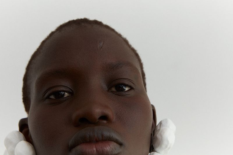 Close-up portrait of an adult female with dark skin, white earrings, and a neutral expression against a bright white background.