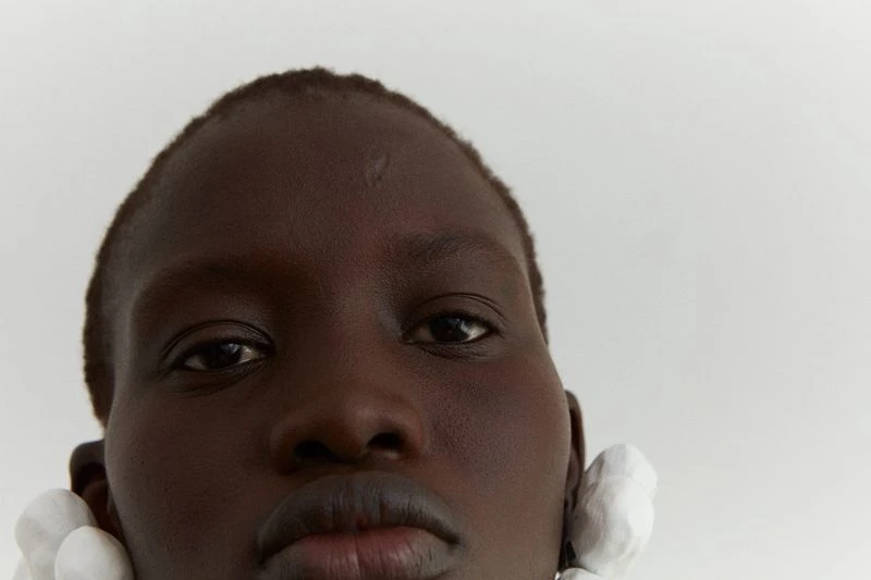 Close-up portrait of an adult female with dark skin, white earrings, and a neutral expression against a bright white background.