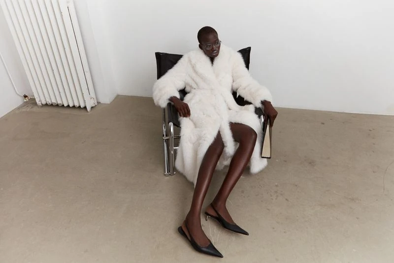 An African-descent female fashion model in a white fur coat and black heels sits in a chair in an indoor photo studio setting.